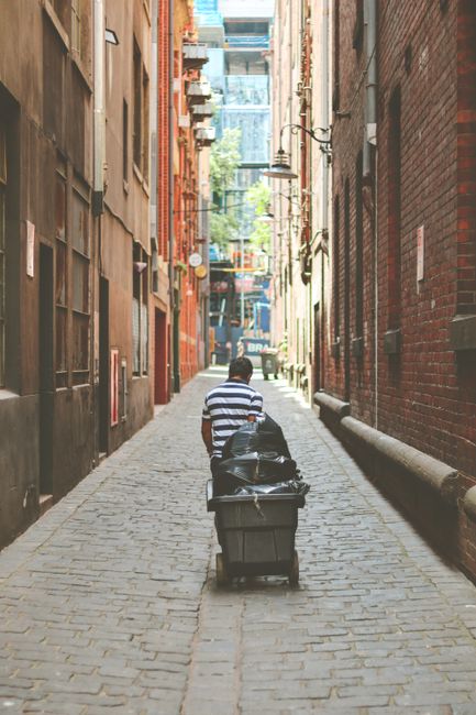 A person wearing a black and white horizontally striped shirt is pushing a large black wheeled waste bin along a narrow cobbled alleyway. The alley is lined with tall brick buildings on both sides, wi