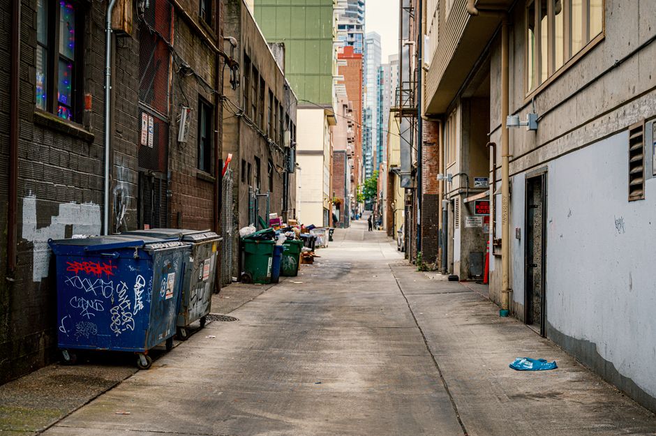 A person wearing a black and white horizontally striped shirt is pushing a large black wheeled waste bin along a narrow cobbled alleyway. The alley is lined with tall brick buildings on both sides, wi