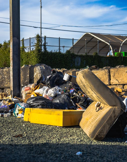 A large blue wheelie bin made of durable plastic with a textured surface, positioned on a paved area beside a building in an outdoor setting. The bin has a black handle on the lid and black markings o